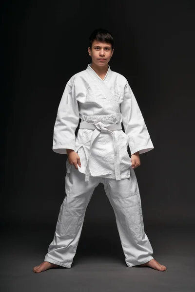 Un adolescent vêtu de vêtements d'arts martiaux pose sur un fond gris foncé, un concept sportif — Photo