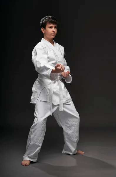 Um adolescente vestido com roupas de artes marciais posa em um fundo cinza escuro, um conceito esportivo — Fotografia de Stock