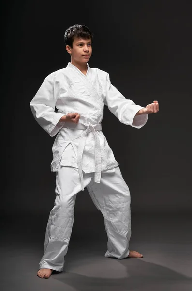 Um adolescente vestido com roupas de artes marciais posa em um fundo cinza escuro, um conceito esportivo — Fotografia de Stock