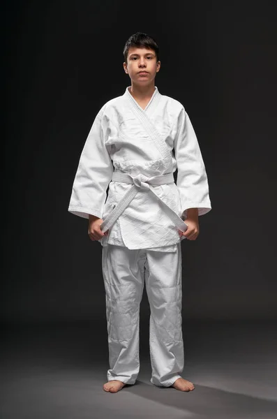 Un adolescente vestido con ropa de artes marciales posa sobre un fondo gris oscuro, un concepto deportivo —  Fotos de Stock