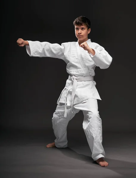 Un adolescente vestido con ropa de artes marciales posa sobre un fondo gris oscuro, un concepto deportivo —  Fotos de Stock