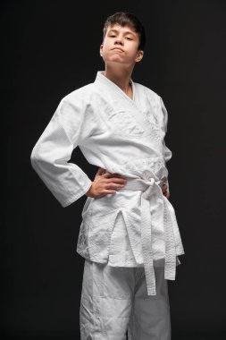 Spor konsepti - dövüş sanatları kıyafeti giymiş gururlu bir genç koyu gri bir arka planda poz veriyor, stüdyo çekimi.