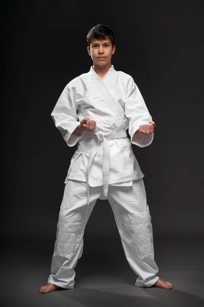 Um adolescente vestido com roupas de artes marciais posa em um fundo cinza escuro, um conceito esportivo — Fotografia de Stock