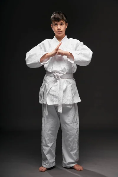 Um adolescente vestido com roupas de artes marciais posa em um fundo cinza escuro, posição de saudação, conceito de esportes — Fotografia de Stock