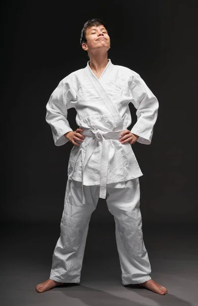 Um adolescente vestido com roupas de artes marciais posa em um fundo cinza escuro, um conceito esportivo — Fotografia de Stock