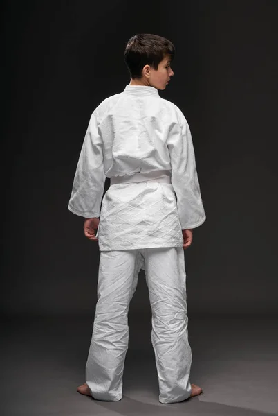 मार्शल आर्ट्स वस्त्रों में पहने हुए एक किशोर का बैकसाइड व्यू एक अंधेरे ग्रे पृष्ठभूमि पर खड़ा है, एक खेल अवधारणा — स्टॉक फ़ोटो, इमेज