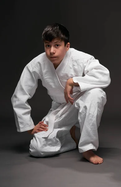 Um adolescente vestido com roupas de artes marciais sentado e pensando em um fundo cinza escuro, um conceito de esportes — Fotografia de Stock