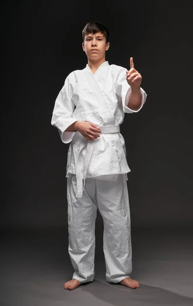 Un adolescente vestido con ropa de artes marciales posando y haciendo gestos sobre un fondo gris oscuro, un concepto deportivo — Foto de Stock
