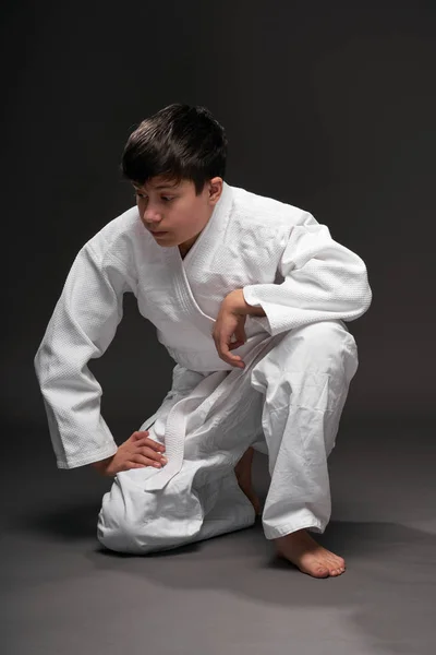 Teenager oblečený v oblečení bojových umění sedí a přemýšlí na tmavě šedém pozadí, sportovní koncept — Stock fotografie