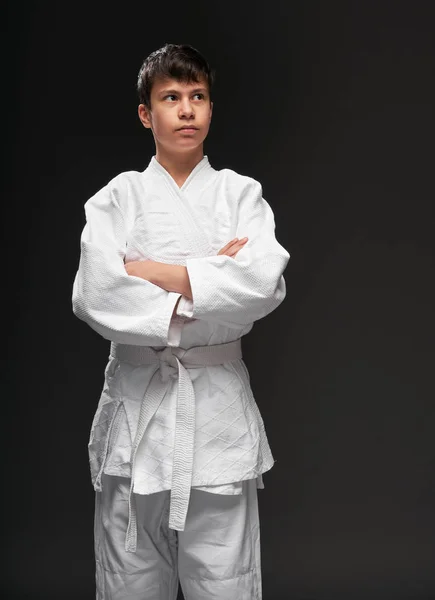 Retrato de un adolescente vestido con ropa de artes marciales posa sobre un fondo gris oscuro, un concepto deportivo —  Fotos de Stock