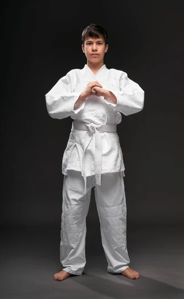 Um adolescente vestido com roupas de artes marciais posa em um fundo cinza escuro, um conceito esportivo — Fotografia de Stock