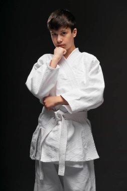 Dövüş sanatları kıyafeti giymiş bir genç koyu gri bir arka planda poz verir.