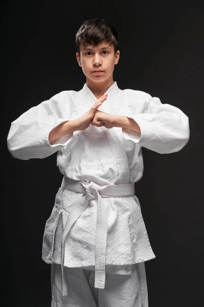 Un adolescente vestido con ropa de artes marciales posa sobre un fondo gris oscuro, posición de saludo, concepto deportivo —  Fotos de Stock