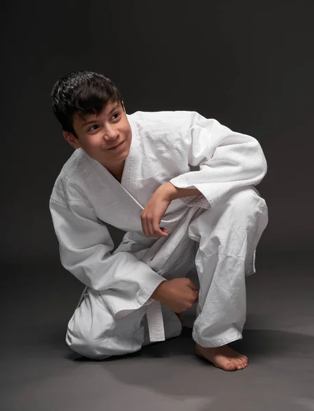 A teenager dressed in martial arts clothing sitting and thinking on a dark gray background, a sports concept — Stock Photo, Image