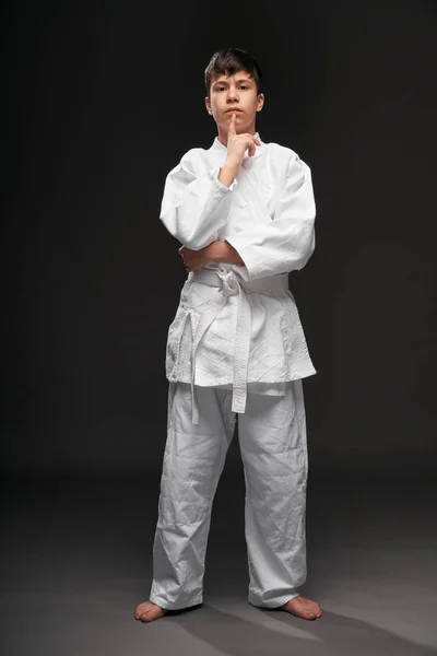 Um adolescente vestido com roupas de artes marciais posa em um fundo cinza escuro, tendo ideia, conceito de esportes — Fotografia de Stock
