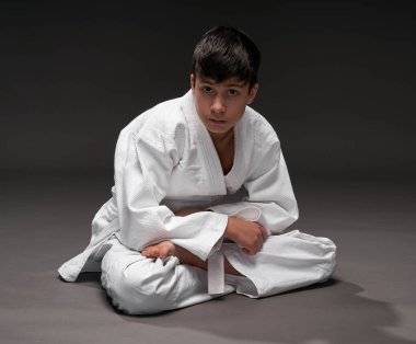 Dövüş sanatları kıyafeti giymiş bir genç koyu gri bir arka planda poz verir.