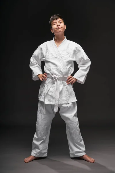 Un adolescente vestido con ropa de artes marciales posa sobre un fondo gris oscuro, un concepto deportivo — Foto de Stock