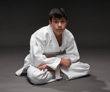 Dövüş sanatları kıyafeti giymiş bir genç koyu gri bir arka planda poz verir.