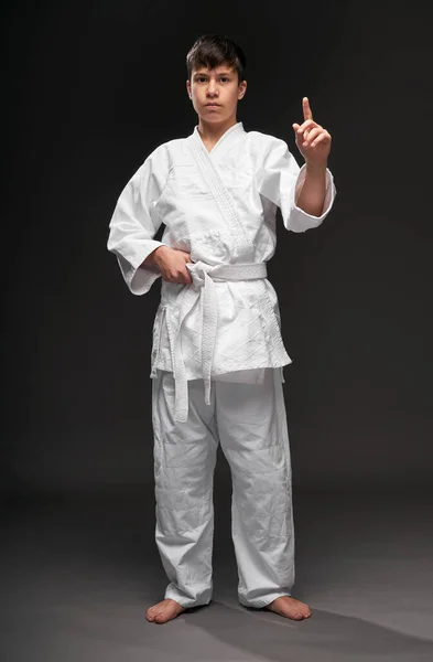 Un adolescente vestito con abiti di arti marziali posa e gesticolando su uno sfondo grigio scuro, un concetto sportivo — Foto Stock