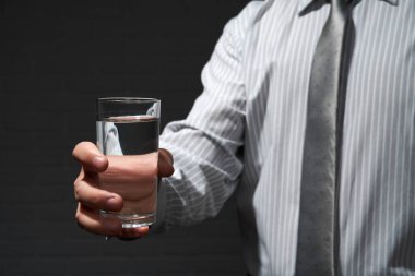 İş adamı yakın plan portresi, bir bardak suyla ayakta duruyor ve poz veriyor, arka planda koyu bir duvar var.