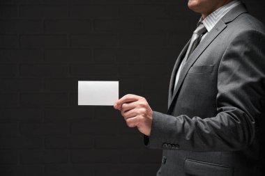 Beyaz, boş bir kartvizit. İş adamı eli, gri takım elbise, koyu duvar arkaplanı.