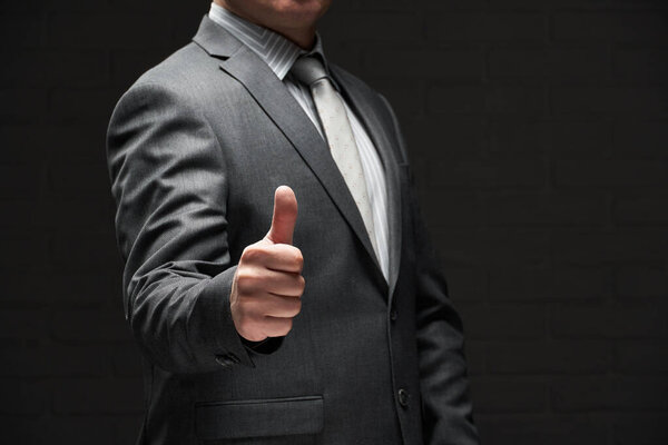 портрет бизнесмена, показывающий лучший жест, большой палец вверх, одетый в серый костюм, темный фон стены
