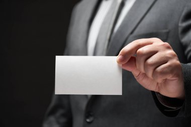 Beyaz, boş bir kartvizit. İş adamı eli, gri takım elbise, koyu duvar arkaplanı.