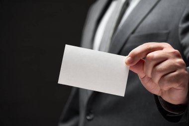 Beyaz, boş bir kartvizit. İş adamı eli, gri takım elbise, koyu duvar arkaplanı.