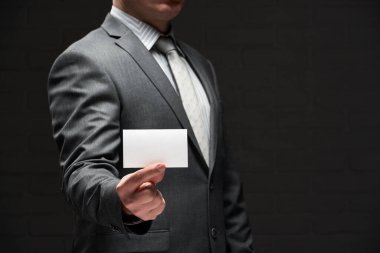 Beyaz, boş bir kartvizit. İş adamı eli, gri takım elbise, koyu duvar arkaplanı.