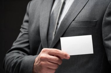 Beyaz, boş bir kartvizit. İş adamı eli, gri takım elbise, koyu duvar arkaplanı.