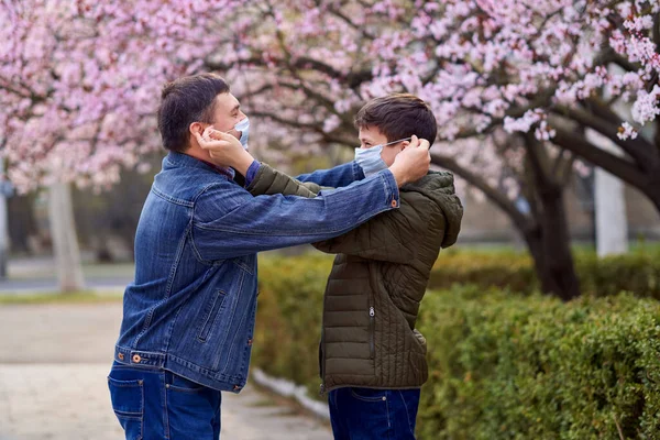 father and son with a face mask are in the city outdoor, blooming trees, spring season, flowering time - concept of allergies and health protection from dusty air