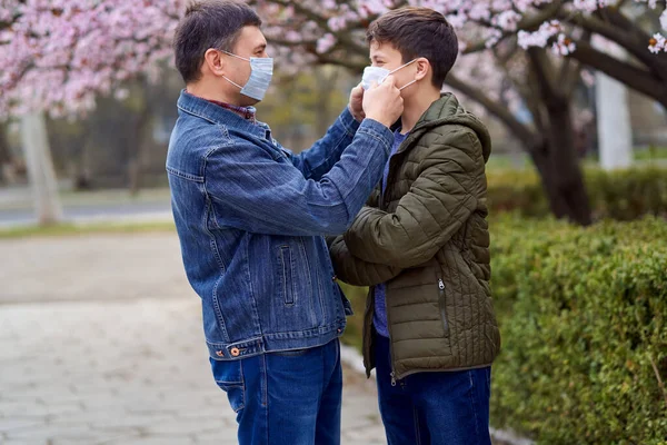顔のマスクを持つ父と息子は街の屋外にあり 木を咲かせ 春の季節 開花期 アレルギーとほこりの多い空気からの健康保護の概念 — ストック写真