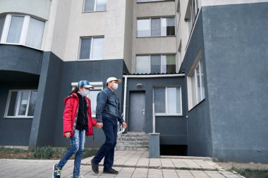 Bir adam ve bir çocuk, apartmanlı yüksek binaların duvarlarının yanında yürüyor. Evleri, kızları, babaları, evleri, yüzlerindeki sağlık maskesi virüslere ve tozlara karşı koruyor.