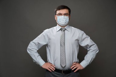 Gri arka planda, tıbbi maske, gözlük, açık çizgili gömlek ve kravat - sıradan ofis kıyafeti, karantina ve antivirüs koruma kavramı - bir iş adamı gibi poz veren bir adam.