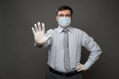 İş adamı gibi giyinmiş bir adam avucunun içi gibi duruyor, gri arka plan, tıbbi maske ve koruyucu eldivenler, gözlük, gömlek ve kravat - karantina ve antivirüs koruması kavramı