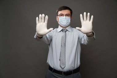 İş adamı gibi giyinmiş bir adam avucunun içi gibi duruyor, gri arka plan, tıbbi maske ve koruyucu eldivenler, gözlük, gömlek ve kravat - karantina ve antivirüs koruması kavramı