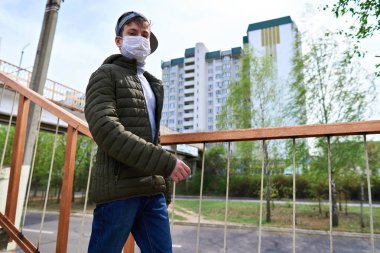 Genç çocuk gün boyunca sokakta yürüyor, yaya geçidi ve apartmanlı yüksek binalar, mesken alanı, yüzünde bir sağlık maskesi virüslere ve toza karşı koruyor.