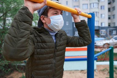 Genç çocuk dışarıda spor malzemelerinin yatay barının yanında duruyor, yüksek katlı binaların yanındaki oyun parkında, yüzünde virüslere ve toza karşı koruyucu bir tıbbi maske var.