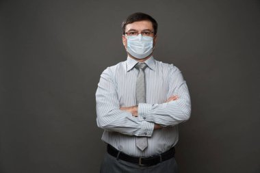 Gri arka planda, tıbbi maske, gözlük, açık çizgili gömlek ve kravat - sıradan ofis kıyafeti, karantina ve antivirüs koruma kavramı - bir iş adamı gibi poz veren bir adam.