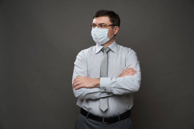 Gri arka planda, tıbbi maske, gözlük, açık çizgili gömlek ve kravat - sıradan ofis kıyafeti, karantina ve antivirüs koruma kavramı - bir iş adamı gibi poz veren bir adam.