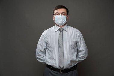 Gri arka planda, tıbbi maske, gözlük, açık çizgili gömlek ve kravat - sıradan ofis kıyafeti, karantina ve antivirüs koruma kavramı - bir iş adamı gibi poz veren bir adam.