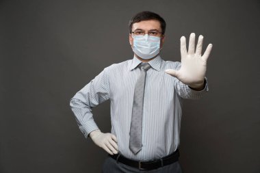 İş adamı gibi giyinmiş bir adam avucunun içi gibi duruyor, gri arka plan, tıbbi maske ve koruyucu eldivenler, gözlük, gömlek ve kravat - karantina ve antivirüs koruması kavramı