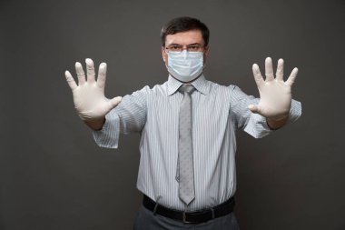 İş adamı gibi giyinmiş bir adam avucunun içi gibi duruyor, gri arka plan, tıbbi maske ve koruyucu eldivenler, gözlük, gömlek ve kravat - karantina ve antivirüs koruması kavramı