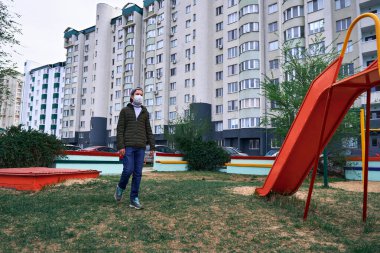 Genç bir çocuk parka doğru yürüyor. Yüksek binaların yanında apartmanlar, yerleşim alanı ve yüzünde virüs ve toza karşı koruyucu bir tıbbi maske var.