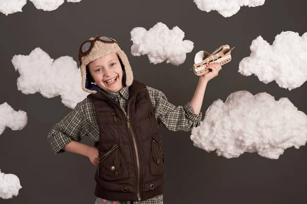a girl plays with a cardboard airplane and dreams of becoming a pilot, dressed in a retro style jacket and helmet with glasses, clouds of cotton wool, gray background, tinted in brown