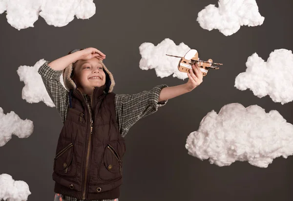 a girl plays with a cardboard airplane and dreams of becoming a pilot, dressed in a retro style jacket and helmet with glasses, clouds of cotton wool, gray background, tinted in brown