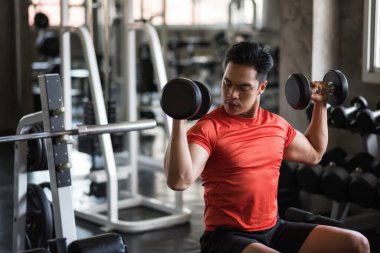 Güçlü Asyalı kaslı adam spor malzemeleri arka planında iki dambılı kullanıyor. Spor fitness merkezinde kaslı vücut sporcusu egzersizi. Vücut geliştirme ve sağlıklı yaşam tarzı konsepti.