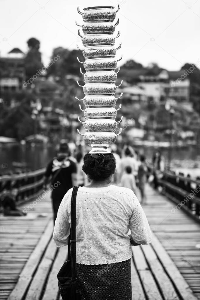 Old woman walk on Mon wooden bridge, the longest in Thailand, with stack of silver pots pile on her head. The tradition and culture in Sangkhlaburi, Kanchanaburi. Black and white photo.