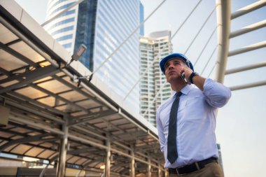 Modern şehirdeki inşaat sahasında kask takan endişeli Amerikalı mühendis. Mimari proje yöneticisi veya kıdemli mühendislik patronu proje gecikmesinden endişe ediyor.