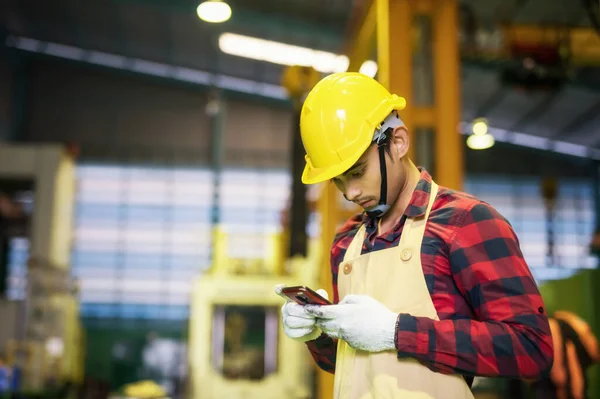 Factory worker with plaid shirt, helmet, safety using  corporate application on smartphone to report work progress to manager. Working at night shift to finish production target. Manufacture industry.